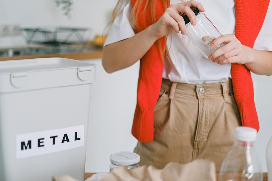 A young woman with long brown hair and light skin stands in a modern kitchen, wearing a white t-shirt and a red vest draped over her shoulders. She is holding a crumpled brown paper bag in her hands, 