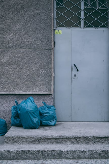 Several blue plastic rubbish bags, filled and tied at the tops, are stacked on the concrete step of an exterior city building entrance. The bags appear to contain household or waste items and are posi