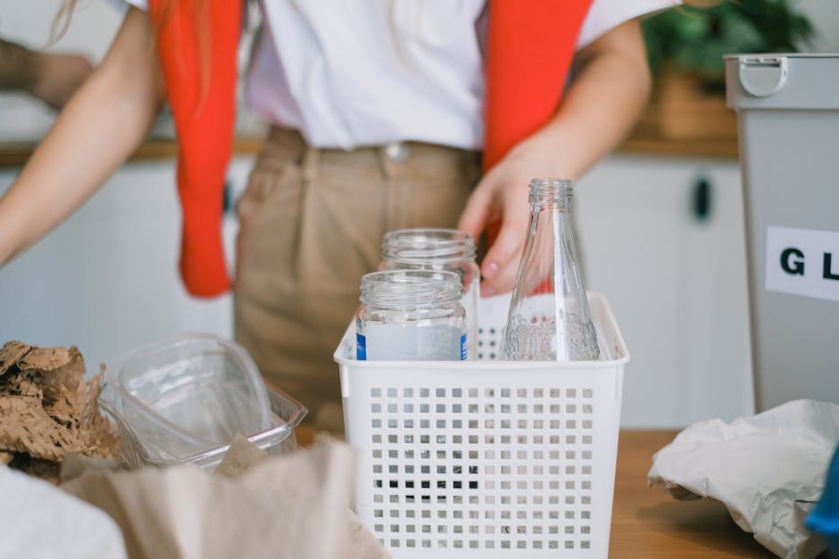 A close-up of a grey plastic waste bin with a white label reading 'PAPER' in black capital letters, positioned on a wooden surface in a domestic interior. The bin is partially filled with crumpled and