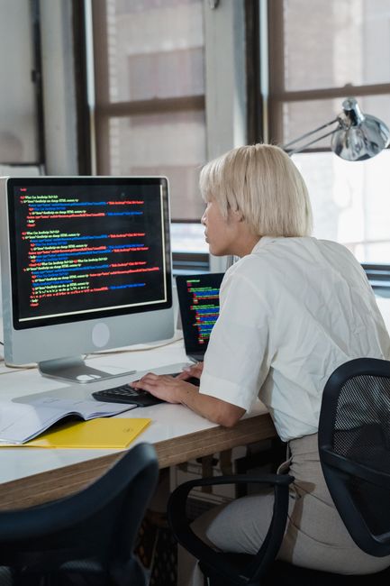 A woman with short blond hair, wearing a white blouse, is seated at a wooden desk in front of a large computer monitor displaying lines of multicoloured code on a black background. She appears to be w